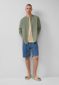 Lichtgroen overhemd met knopen over een beige t-shirt, blauwe denim shorts en beige instap sandalen. Model staat met de handen in de zakken.