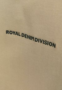 Bezs tekstila ar izšūtu tekstu "ROYAL DENIM DIVISION" melnā krāsā. Audumam ir gluda, vienmērīga tekstūra.