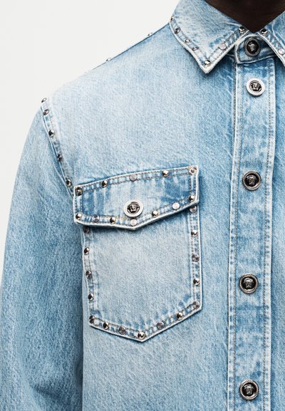 Φωτεινό μπλε τζιν μπουφάν με ασημένιες τρουκς που περιγράμμιζουν την τσέπη, τον ώμο, το γιακά και μοναδικά ασημένια κουμπιά με κεφάλι Μέδουσας.