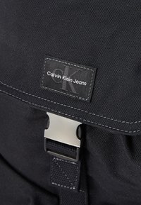 Svart stoffryggsekk med en sølvfarget metallspenne og sydd logo-merke med "Calvin Klein Jeans." Teksturert materiale med slitesterk konstruksjon.