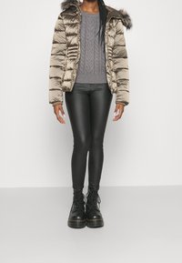 Doudoune beige métallisé avec une capuche bordée de fourrure, associée à un pull en maille torsadée gris et un legging en similicuir noir. Bottes noires.