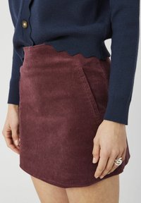 Jupe en velours côtelé bordeaux avec une taille festonnée et des poches latérales. La jupe a une texture lisse et une coupe ajustée, associée à un pull marine.