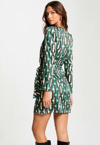 Robe à manches longues en tissu satiné, avec des motifs ovales verts, bruns et blancs. Elle dispose d'une taille nouée et d'une silhouette courte.