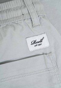 Pantalones gris claro con una cinturilla elástica, una etiqueta cosida con "Reell" y "est. 1997", y un detallado acolchado en la tela.