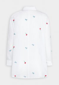 Chemise blanche à manches longues, ornée d'une broderie florale colorée en rouge et bleu. Les détails incluent un col et des poignets boutonnés.