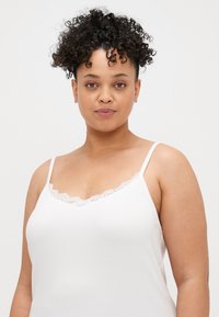 Camisola branca com alças finas em espaguete, apresentando um acabamento de renda ao longo da gola, feita de um tecido ribflat com uma textura suave.