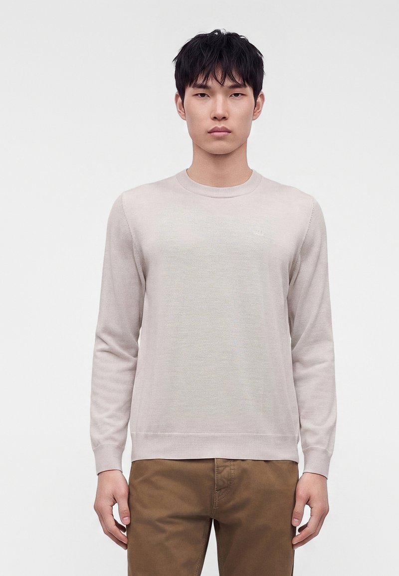 Jeune homme debout, face à l’avant, portant un pull beige clair à manches longues et un pantalon marron sur un fond blanc uni.