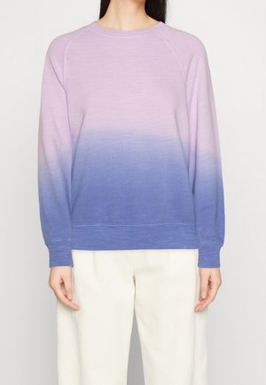 Kvinde iført en langærmet sweatshirt med rund halsudskæring med pink til blå ombré-design og hvide bukser, stående mod en ensfarvet baggrund.