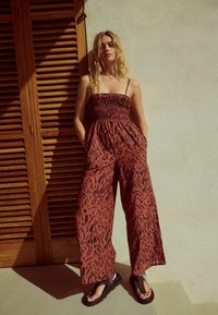 Röd-brun jumpsuit med ett rynkat liv, vida ben och svarta abstrakta mönster. Kombinerad med svarta sandaler med en tåslingsdesign.