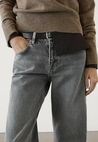 Jeans in denim grigio con una vestibilità rilassata, abbinati a una cintura nera e a un maglione a coste marrone, che mostrano materiali testurizzati e un design a strati.