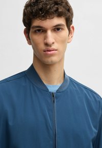Blaue Bomberjacke mit glatter, matter Textur, geripptem Kragen und Reißverschluss vorne. Getragen über einem hellblauen Hemd.