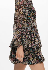 Blommig klänning med svart genomskinligt tyg, volangärmar, smockad midja och lager på nederkanten. Har flerfärgade blommor och blad. Silverringar på handen.