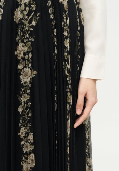 Χέρι που ακουμπά δίπλα σε μαύρη πλισέ φούστα με κάθετα μπεζ άνθη, σε συνδυασμό με μακρυμάνικο λευκό πουκάμισο με ορατό μανίκι.