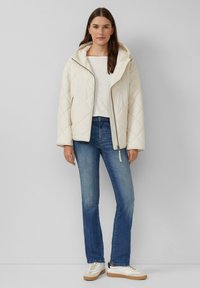 Gepolsterte Cremejacke mit Kapuze, Reißverschluss und Seitentaschen, kombiniert mit blauen Jeans und weißen Sneakers mit beigen Sohlen.