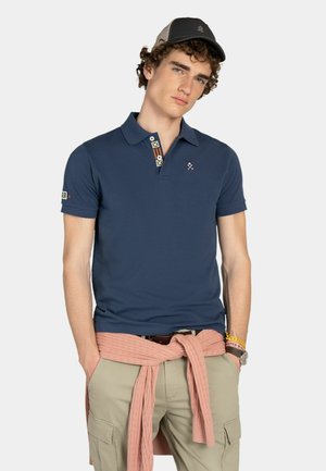 Joven con camiseta tipo polo azul marino, pantalones cargo beige, suéter rosa atado alrededor de la cintura, gorra negra y gris y pulseras, de pie con las manos en los bolsillos.