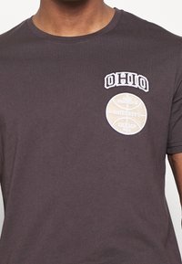 T-shirt gris foncé avec le texte « OHIO » et un patch graphique beige représentant un ballon de basketball portant l'inscription « Ohio Brooklyn University Champ 2014 » sur la poitrine.