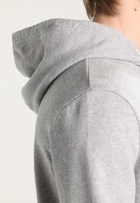 Primer plano de una persona que lleva una sudadera ligera gris claro, mostrando detalles de la capucha y la costura del hombro.