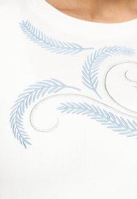 Λευκό ύφασμα με ανοιχτό γαλάζια κεντημένα φύλλα που μοιάζουν με φτέρη και ασημένιες στροβιλιζόμενες γραμμές που σχηματίζουν ένα διακοσμητικό μοτίβο κοντά σε γραμμή λαιμού.