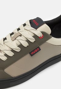 Sneaker features olive green and beige materials, textured surface, beige laces, red accents, and a black logo patch on the tongue.