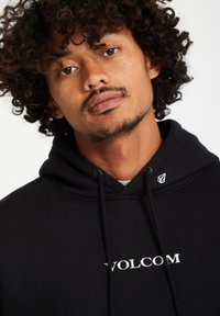 Čierna katoenová mikina s kapucňou a bielym logom "VOLCOM" na prednej strane, so šnúrkou na kapucni a malým bielym emblémom na pleci.