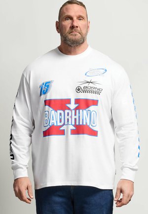 Mand med kort hår og skæg iført hvid skjorte med lange ærmer med rød og blå "BADRHINO" logo og racer-stil grafiske print, står stille.