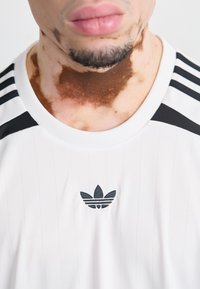 White athletic shirt with vertical pinstripes, black accents on shoulders, and a black embroidered logo on the chest.