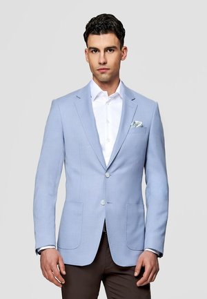 Mężczyzna w jasnoniebieskiej marynarce, białej koszuli i ciemnobrązowych spodniach, stojący na jednolitym szarym tle.