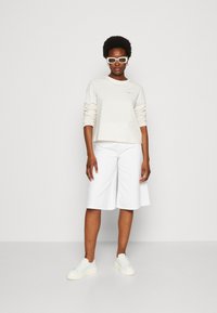 Maglietta a maniche lunghe crema con maniche arricciate, abbinata a pantaloni corti bianchi fino al ginocchio e sneakers bianche. Gli occhiali da sole bianchi completano il look.