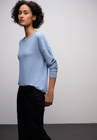 Světlomodrý svetr s volným střihem, kulatým výstřihem a žebrovanými manžetami, kombinovaný s tmavými džínami na jednoduchém pozadí.