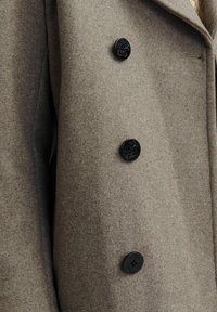 Manteau en laine marron avec trois boutons noirs ornés de motifs en relief. Le tissu a un aspect doux et texturé, mettant en valeur sa coupe sur mesure.