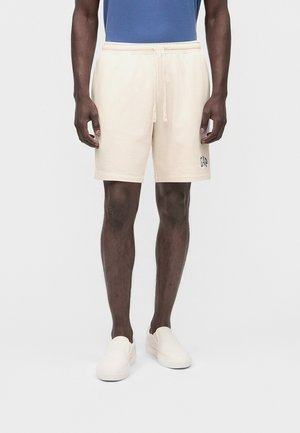 Férfi, aki bézs zsinóros rövidnadrágot visel "GAP" logóval és fehér csúszásmentes cipőben áll, egy egyszínű világos háttér előtt.