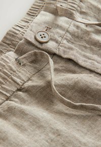 Tela de lino marrón claro con textura visible, que presenta un botón texturizado y un lazo de tela. El diseño incluye una cinturilla elástica.