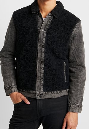 Person trägt eine schwarze Sherpa-Weste über einer ausgebleichten schwarzen Jeansjacke mit Knopfleiste, eine Hand in der Tasche, vor weißem Hintergrund.