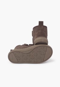 Botines de ante marrón con suela de goma texturizada. Incluyen una lengüeta trasera para tirar y correas ajustables para un ajuste seguro.