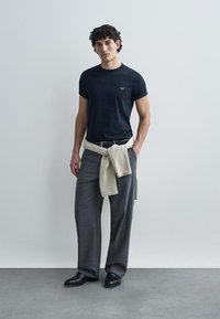 Tamsiai mėlynas aptemptas marškinėlis su mažu logotipu, šviesiai smėlio spalvos megztinis rišamas aplink juosmenį, pilkos, plačios kelnės ir juodi batai.