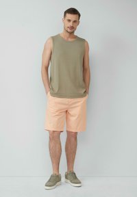 Olivgrünes ärmelloses Oberteil, pfirsichfarbene Shorts und graue Sneakers. Die Shorts haben eine glatte Textur; das Oberteil ist gerippt, figurbetont und lässig.