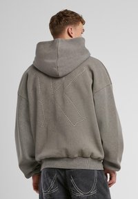 Gray hoodie with a large, stitched letter "K" on the back. Features a drawstring hood and ribbed hem. Fabric has a soft texture.