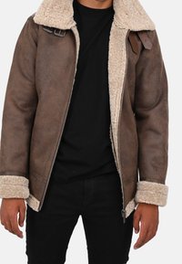 Hnědá ovčí bunda s krémovým umělým kožíškem na límci a manžetách, s předním zipem a nastavitelnými popruhy na ramenou.