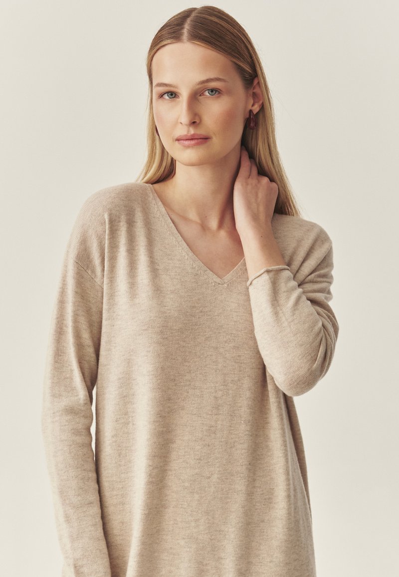 Beżowy sweter w serek z długimi rękawami, wykonany z miękkiego materiału; charakteryzuje się luźnym krojem i delikatną teksturą. Brak wyraźnych wzorów lub akcentów.