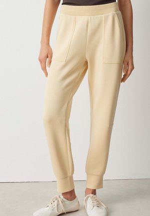 Femme portant un pantalon de jogging en maille beige avec taille élastique et poches appliquées, associé à des baskets blanches à lacets.