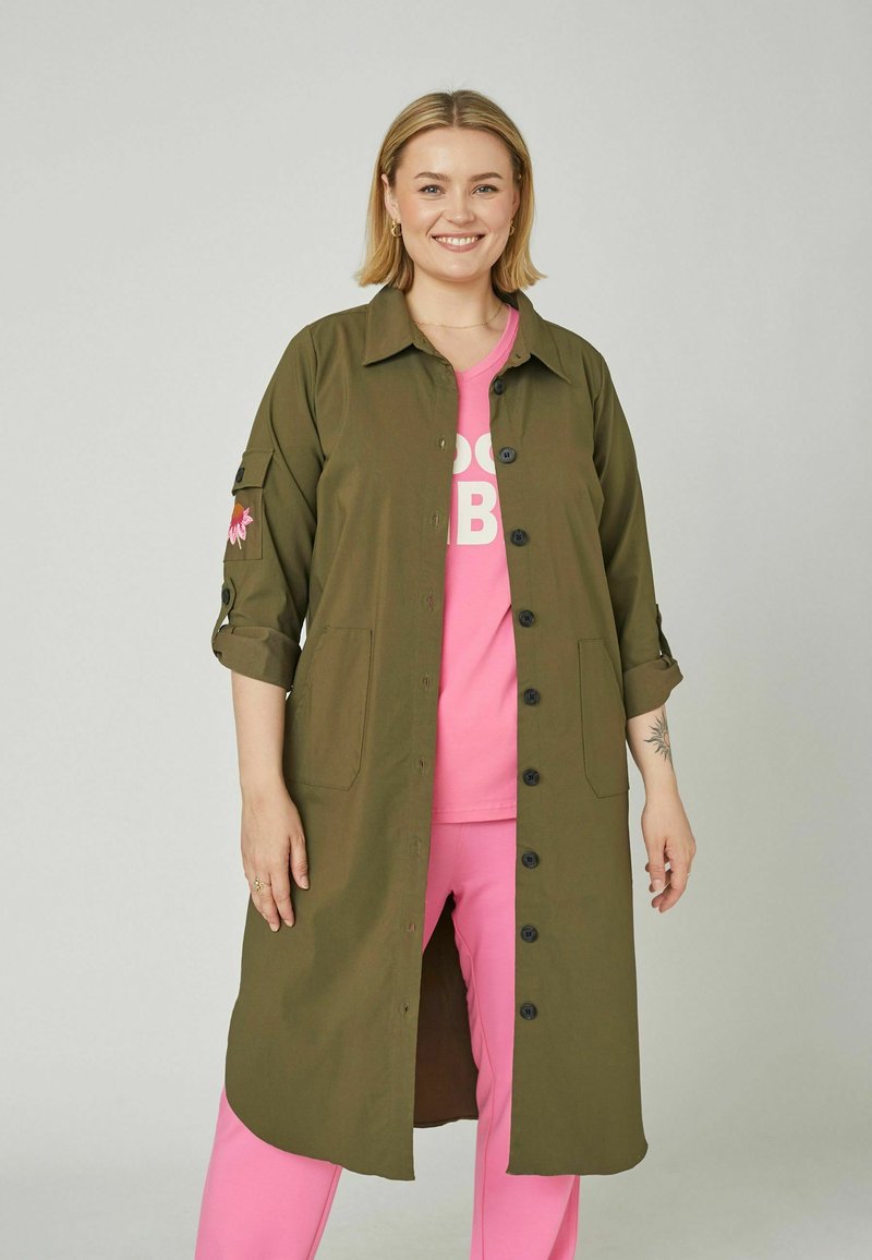 Femme souriante aux cheveux blonds portant un long manteau boutonné vert olive sur une chemise et un pantalon rose vif, debout devant un fond gris uni.