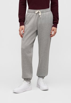 Personne portant un pantalon de survêtement gris avec cordon à la taille, des baskets blanches et un haut à manches longues rouge foncé sur un fond uni.