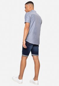 Lichtblauw overhemd met korte mouwen, denim shorts met opgerolde zomen en witte sneakers. Materiaal lijkt glad; het overhemd heeft een kraag.