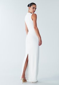 Robe blanche sans manches avec une silhouette ajustée, une fente latérale et un détail de fronces à l'arrière. Le mannequin porte des talons dorés et des accessoires minimalistes.