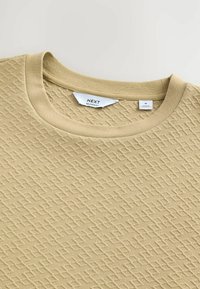 Camisa de manga corta beige claro con una tela texturizada de patrón de diamante. Presenta un cuello redondo acanalado. Etiqueta de la marca visible en el interior.