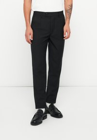 Schwarze Hosen mit maßgeschneidertem Schnitt, ausgestattet mit einem Knopfverschluss, seitlichen Taschen und einer glatten Stofftextur. Getragen mit schwarzen Slippern.