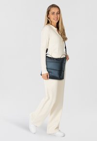 Bolso bandolera acolchado en color marino con un acabado texturizado, que cuenta con una larga correa ajustable, llevado por una persona vestida con un conjunto blanco.