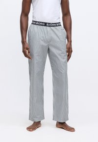 Vīrietis valkā melnbaltas vertikālas svītrainas pidžamu bikses ar melnu elastīgu jostu, zīmols "BJÖRN BORG", stāvot basām kājām.