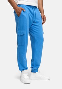 Blaue Baumwoll-Jogginghose mit Seitentaschen, elastischem Bund und Bündchen an den Knöcheln. Tragen mit weißen Sneakern. Verfügt über eine Cargo-Tasche an der Seite.
