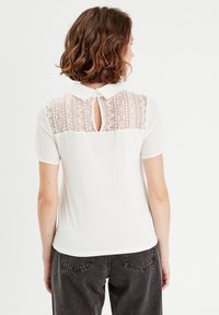 Weißes Poloshirt mit einem Spitzenrückenpanel, das über ein Schlüssellochdetail und kurze Ärmel verfügt. Weicher Stoff mit zarten Mustern auf der Spitze.